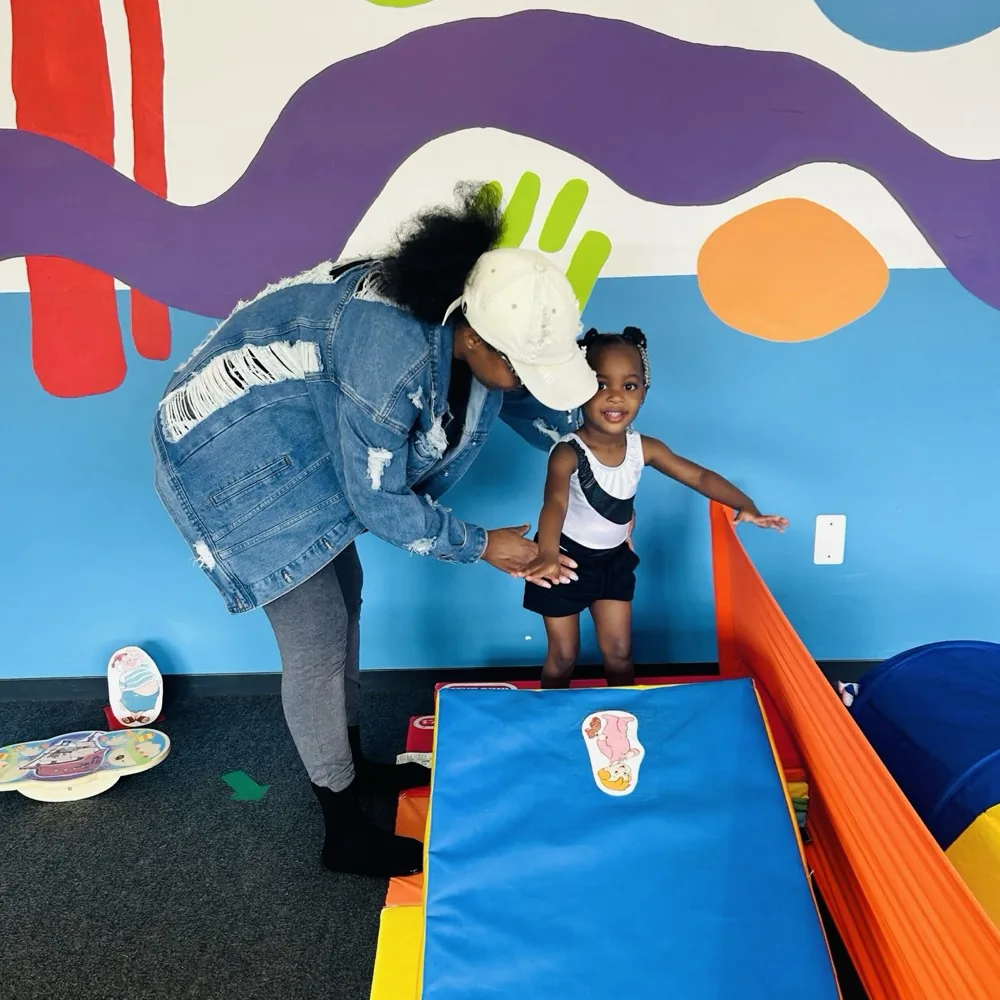 A caregiver helps a smiling toddler balance on a soft wedge in the colorful preschool gym