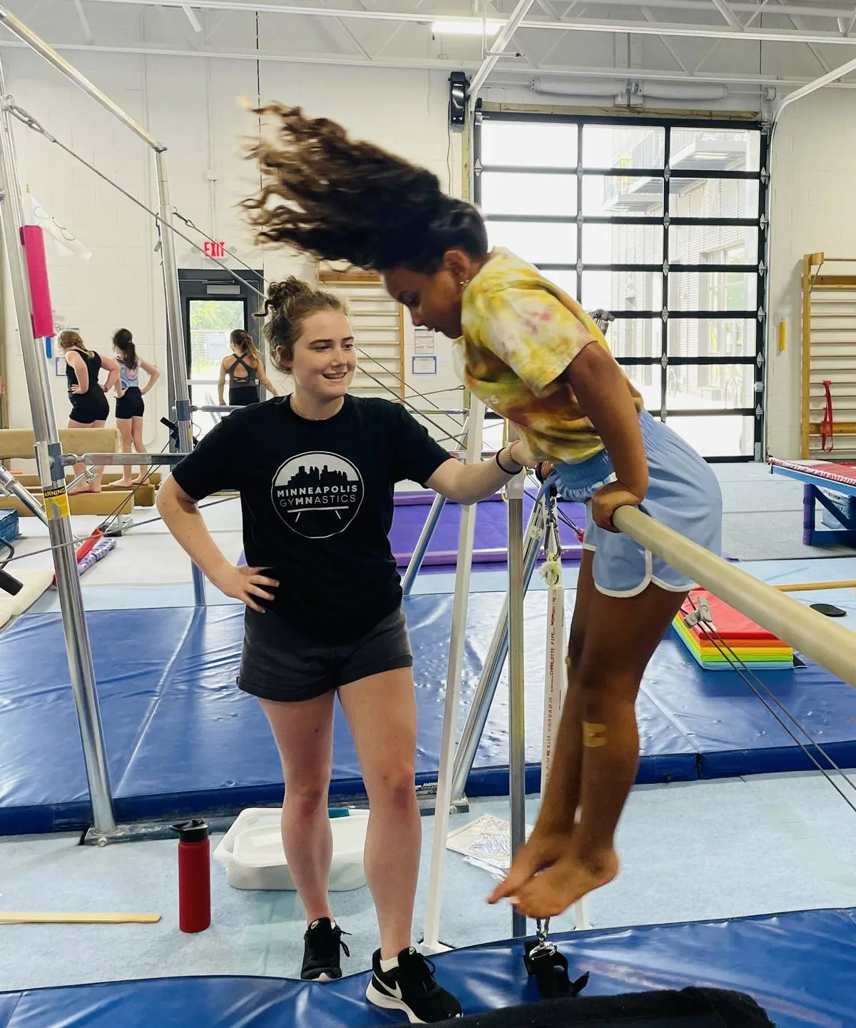 A smiling coach spots a young gymnast swinging up onto the uneven bars